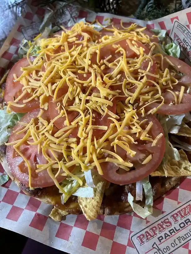 A close-up photo of a loaded pizza or nacho dish topped with shredded cheddar cheese, thick tomato slices, and lettuce…