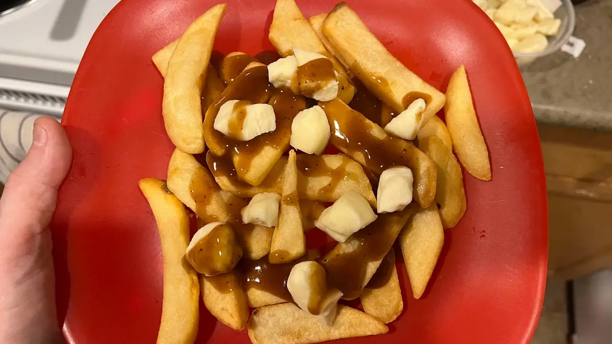 A hand holds a red plate filled with golden-brown french fries topped with brown gravy and white cheese curds. The image is…
