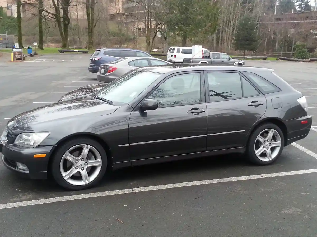 A dark gray 2003-2005 Lexus IS300 sedan is parked in an outdoor lot, its sleek profile and silver alloy wheels visible. The…