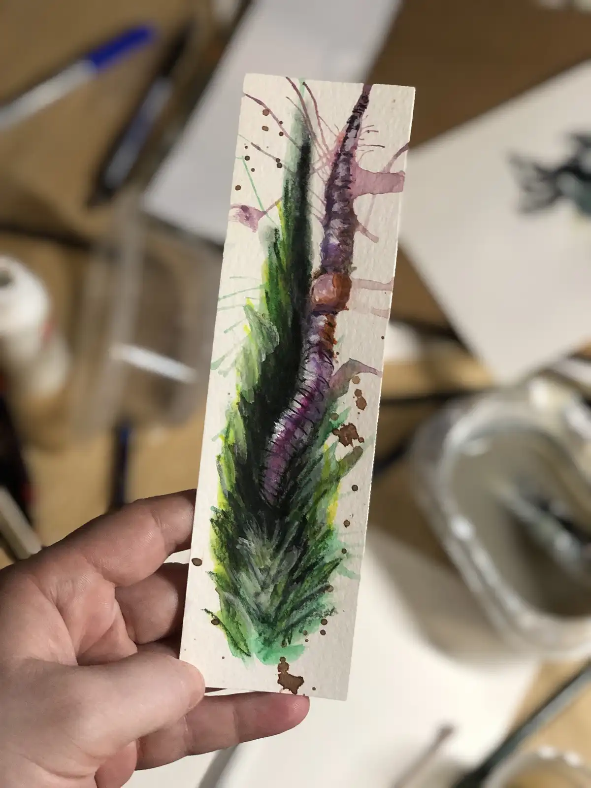 A hand holds up a small rectangular watercolor painting depicting a green leafy plant with a purple root, resembling a…