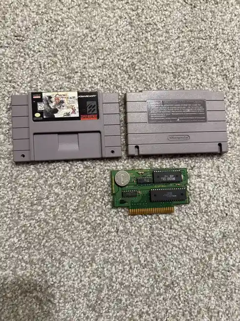 Two gray Super Nintendo game cartridges lie on a carpet, one showing the 'Chrono Trigger' label and the other its plain…