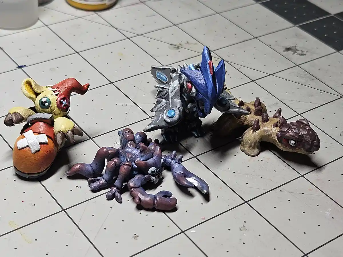 Four handmade clay monster figurines are arranged on a tiled floor, one with a bandaged head and orange body, another with…