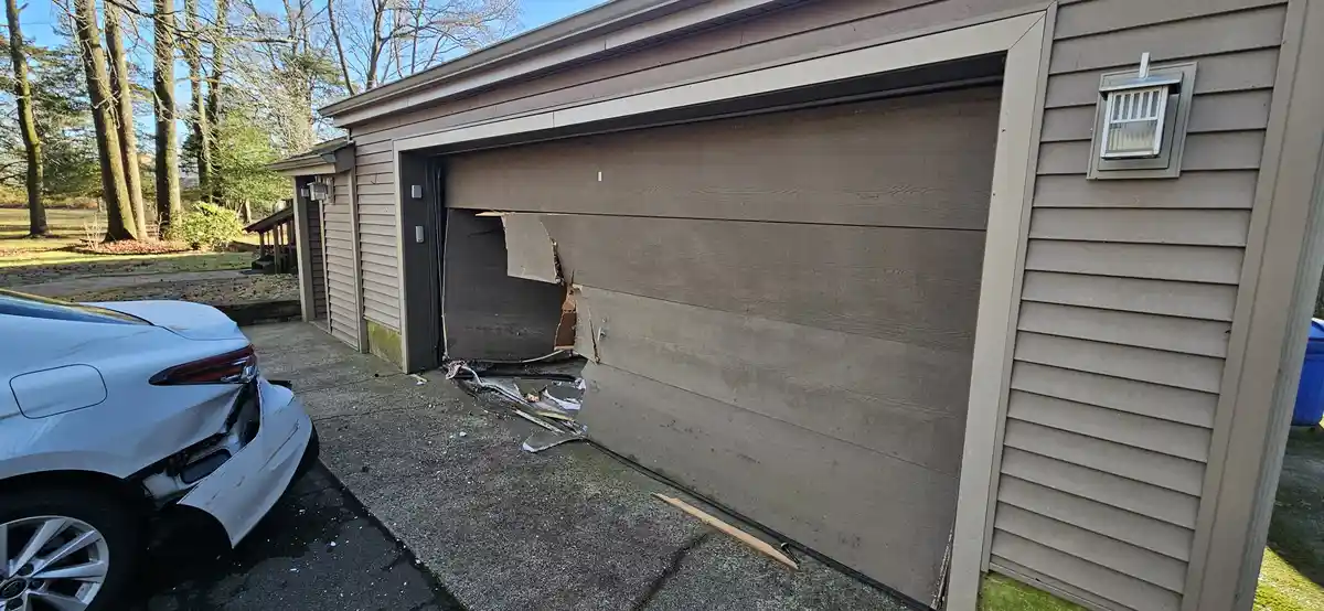 A white car with minor rear damage is parked next to a severely damaged garage door that has a large, jagged hole punched…