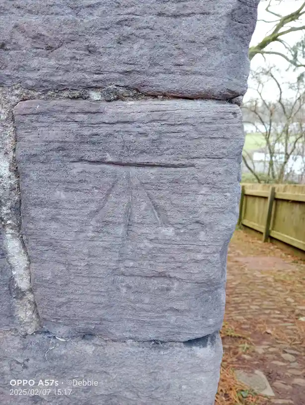 A close-up photo shows a rough, grey stone wall with a crudely carved face etched into one of the stones. The face has two…