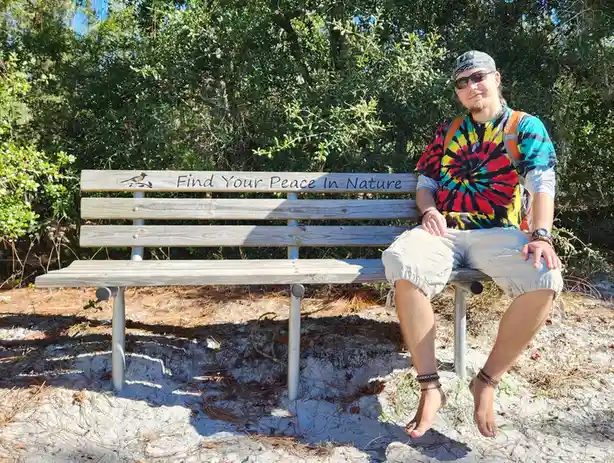 A man in a colorful tie-dye shirt, bandana, and sunglasses sits barefoot on a wooden park bench engraved with the phrase…
