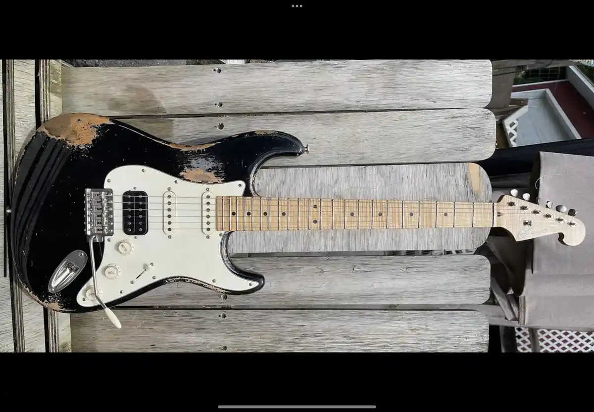A black electric guitar with a heavily worn, chipped finish lies on a weathered wooden bench. The guitar has a white…