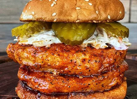 A close-up photo of a burger with two stacked, saucy chicken tenders, topped with pickles and coleslaw, all between a sesame…