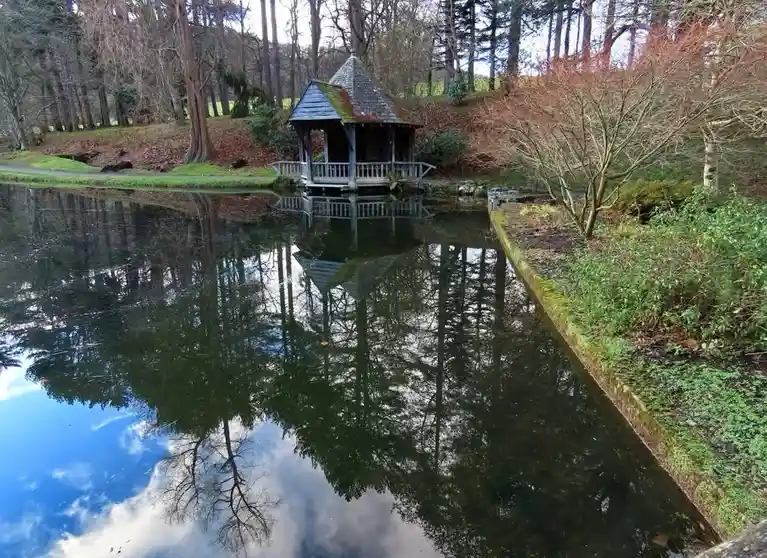 A serene, still pond reflects a wooden gazebo and surrounding trees, with a clear blue sky and clouds mirrored perfectly in…