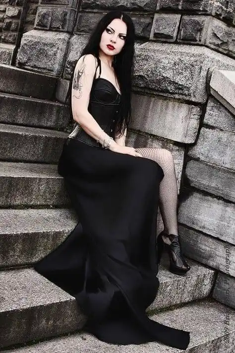 A woman with long black hair, heavy makeup, and a tattoo on her left arm sits on stone steps against a stone wall. She wears…