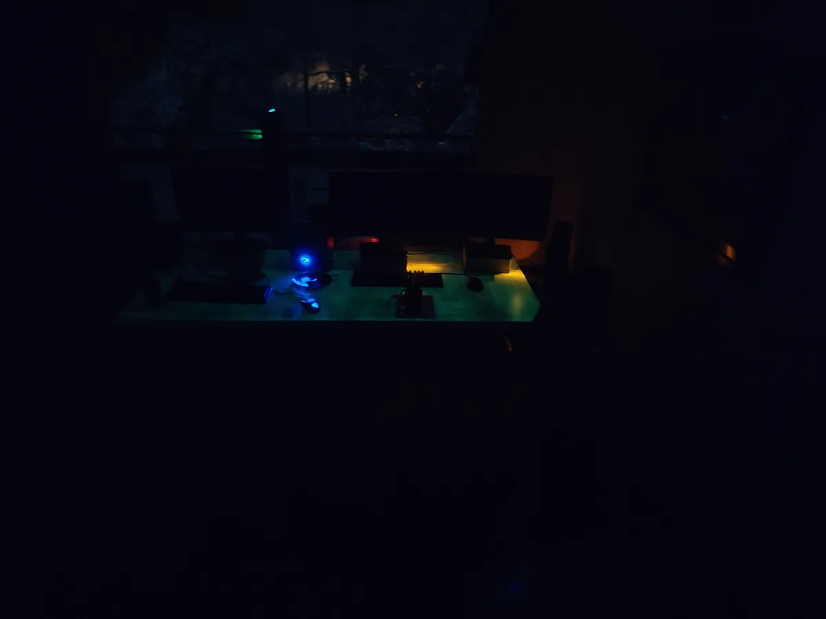 A dimly lit room with a computer desk setup illuminated by blue and yellow RGB lighting from peripherals, featuring two…