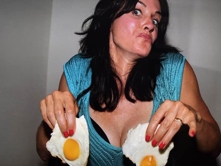 a woman with dark hair and red fingernails holds two fried eggs, one in each hand, looking directly at the camera with a…