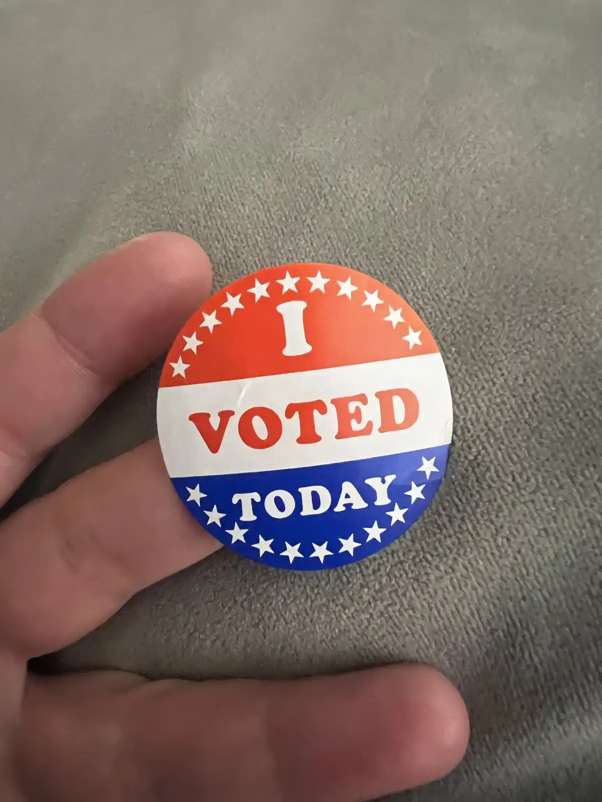 A hand holds a circular red, white, and blue 'I VOTED TODAY' button against a gray textured background. The button features…
