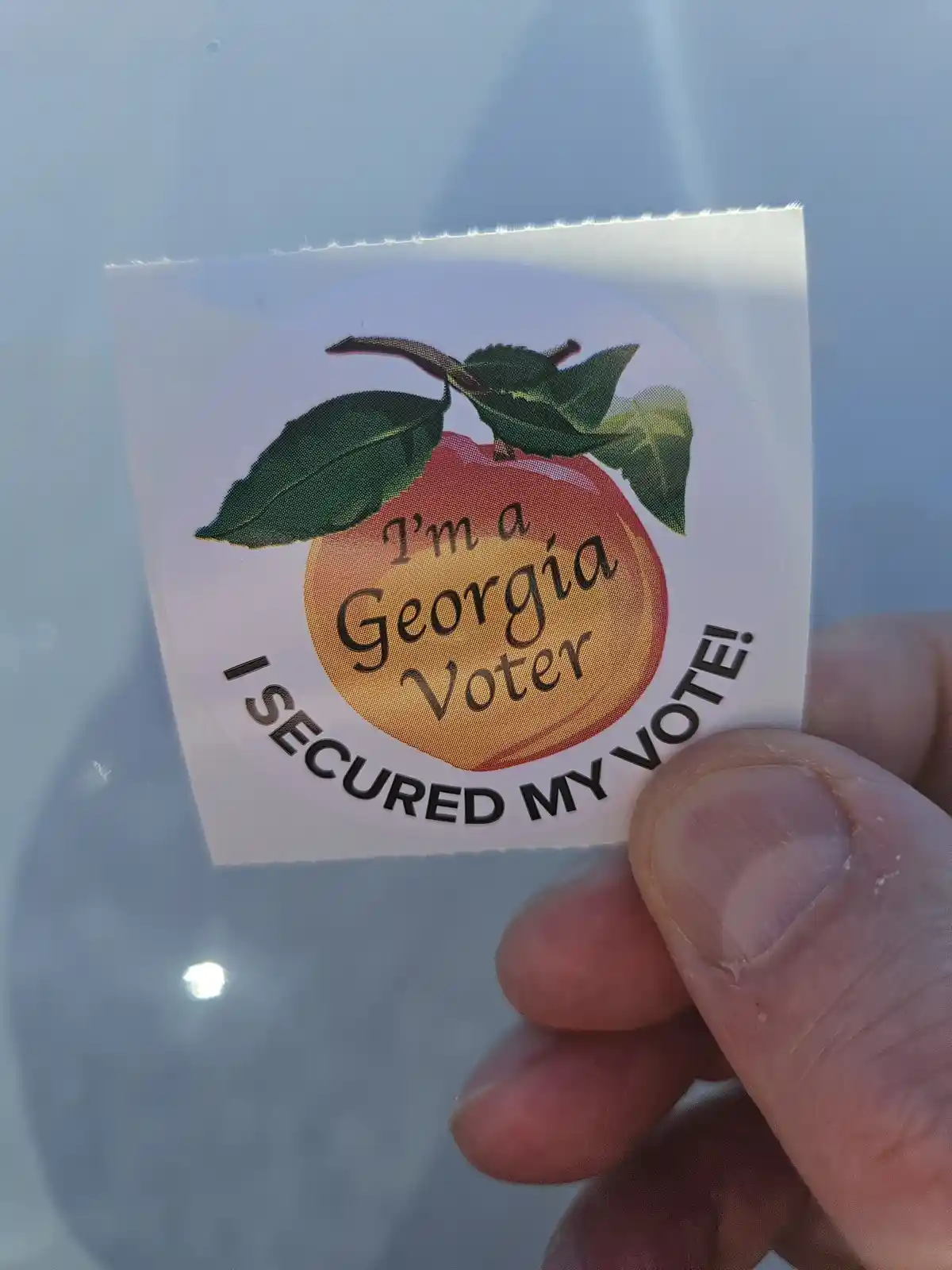 A hand holds a circular sticker featuring a peach with green leaves and the text 'I'm a Georgia Voter' inside the fruit…
