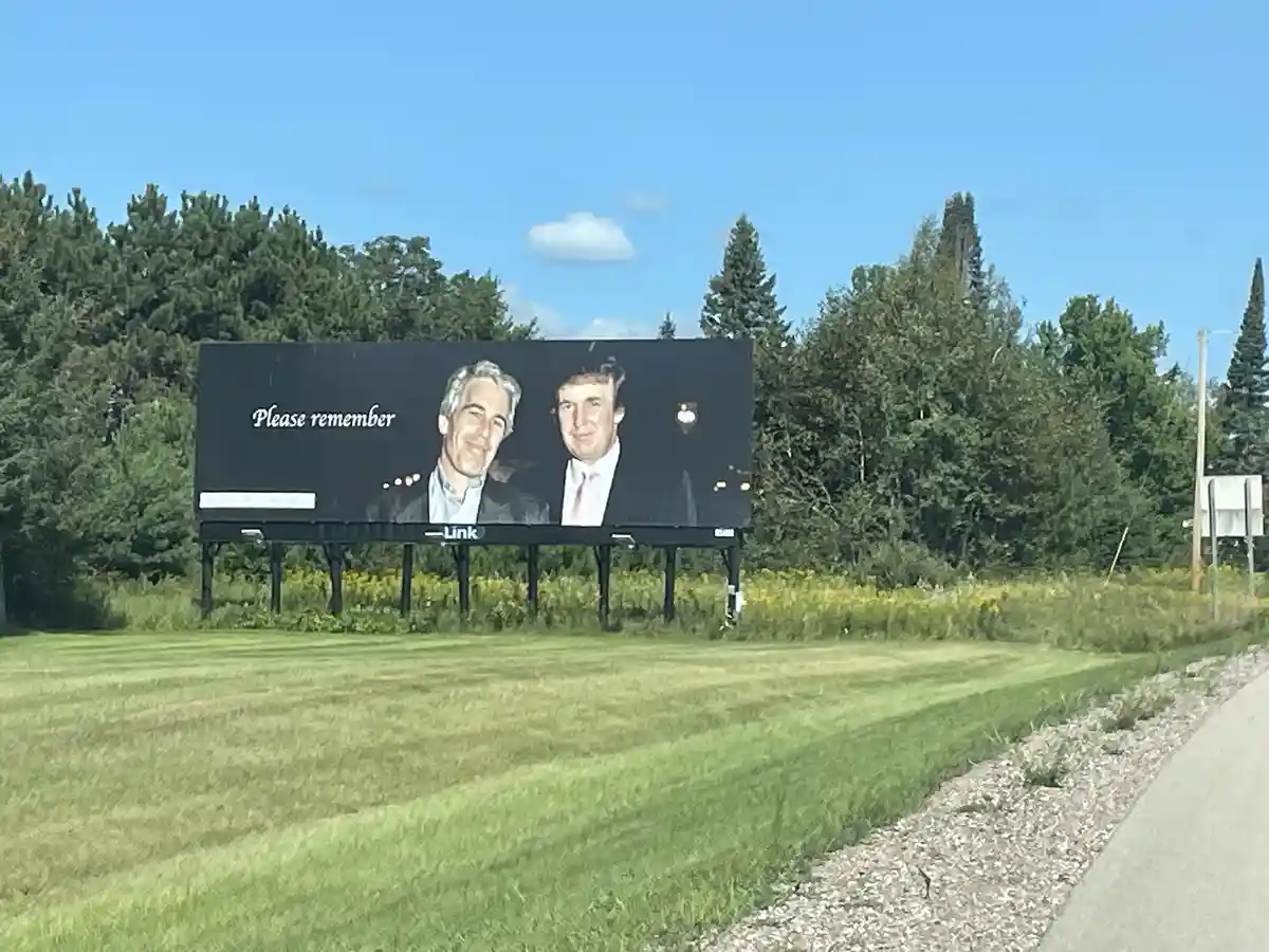 A large outdoor billboard displays a photo of two men, one with gray hair and one with dark hair, smiling side by side…