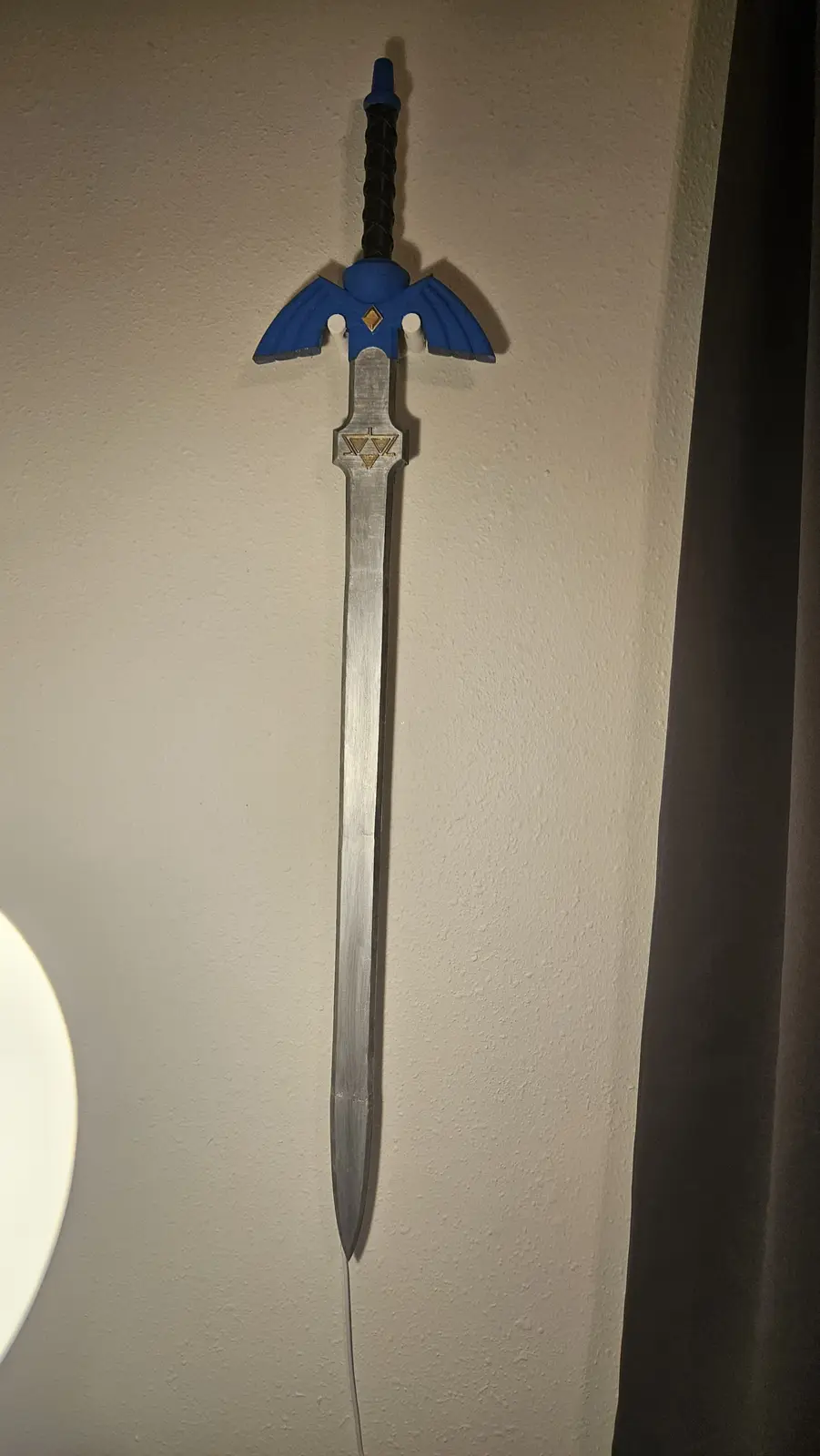 A replica of the Master Sword from The Legend of Zelda hangs vertically on a beige wall, its silver blade and blue hilt with…