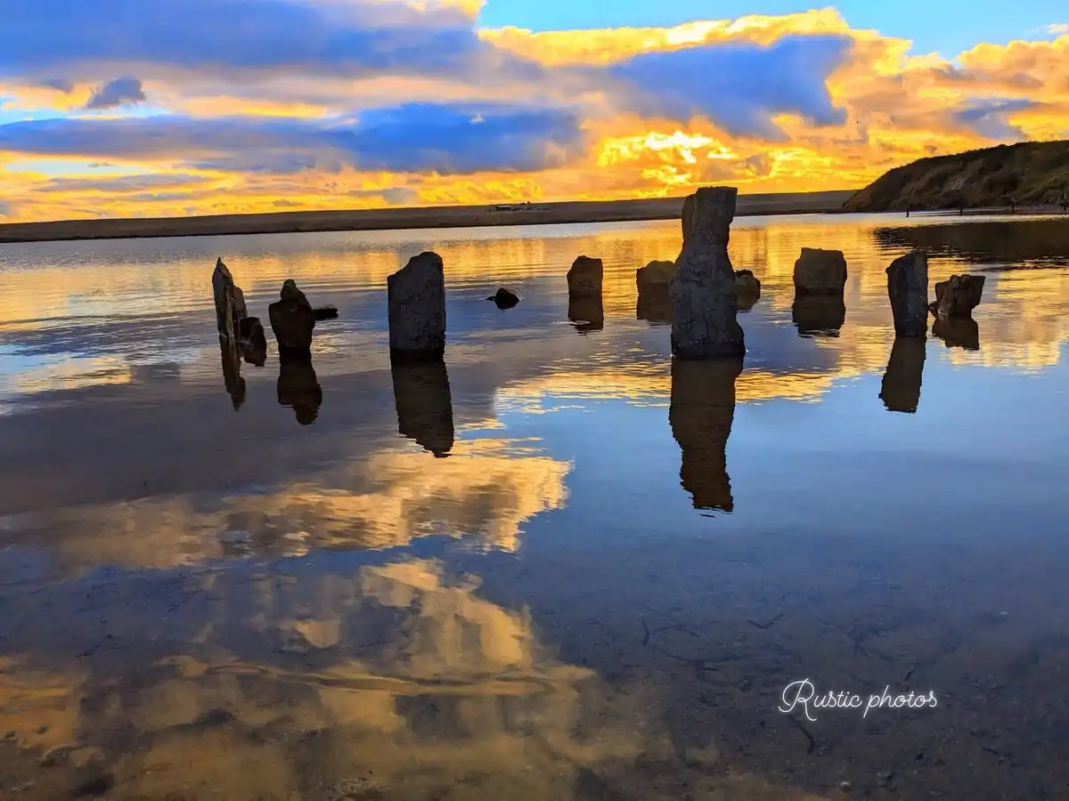 A serene landscape photo shows several large, weathered stone pillars standing in shallow water, reflecting the vibrant…