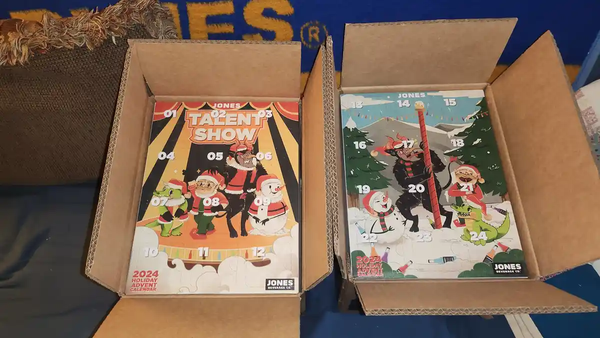 Two open cardboard boxes display Jones Beverage Co. 2024 Holiday Advent Calendars, each featuring cartoon characters in…