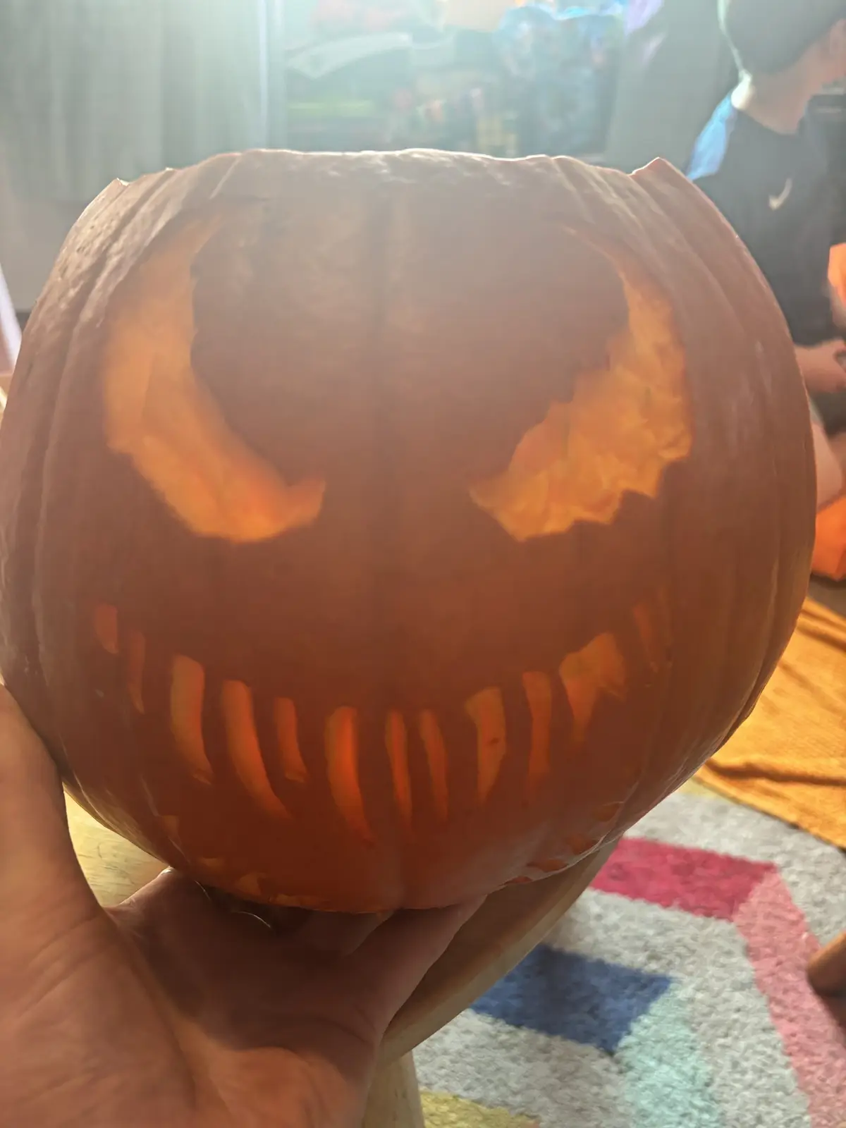 A hand holds a carved Halloween pumpkin with a menacing face, featuring jagged, uneven eyes and a wide, toothy grin. The…