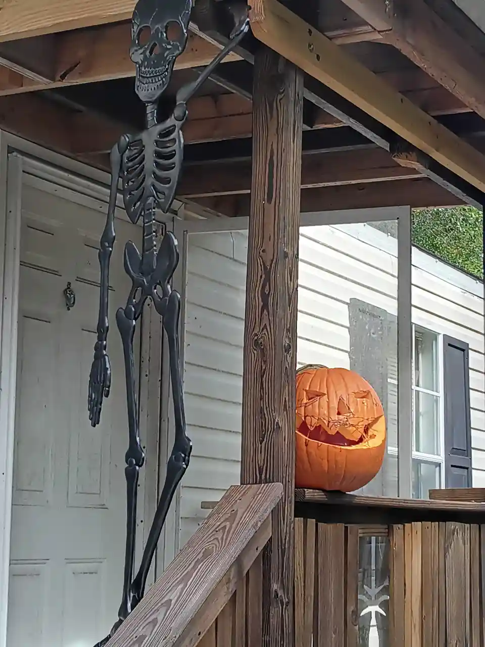A black plastic skeleton decoration hangs from a porch roof beam next to a white door, while a carved jack-o'-lantern with a…