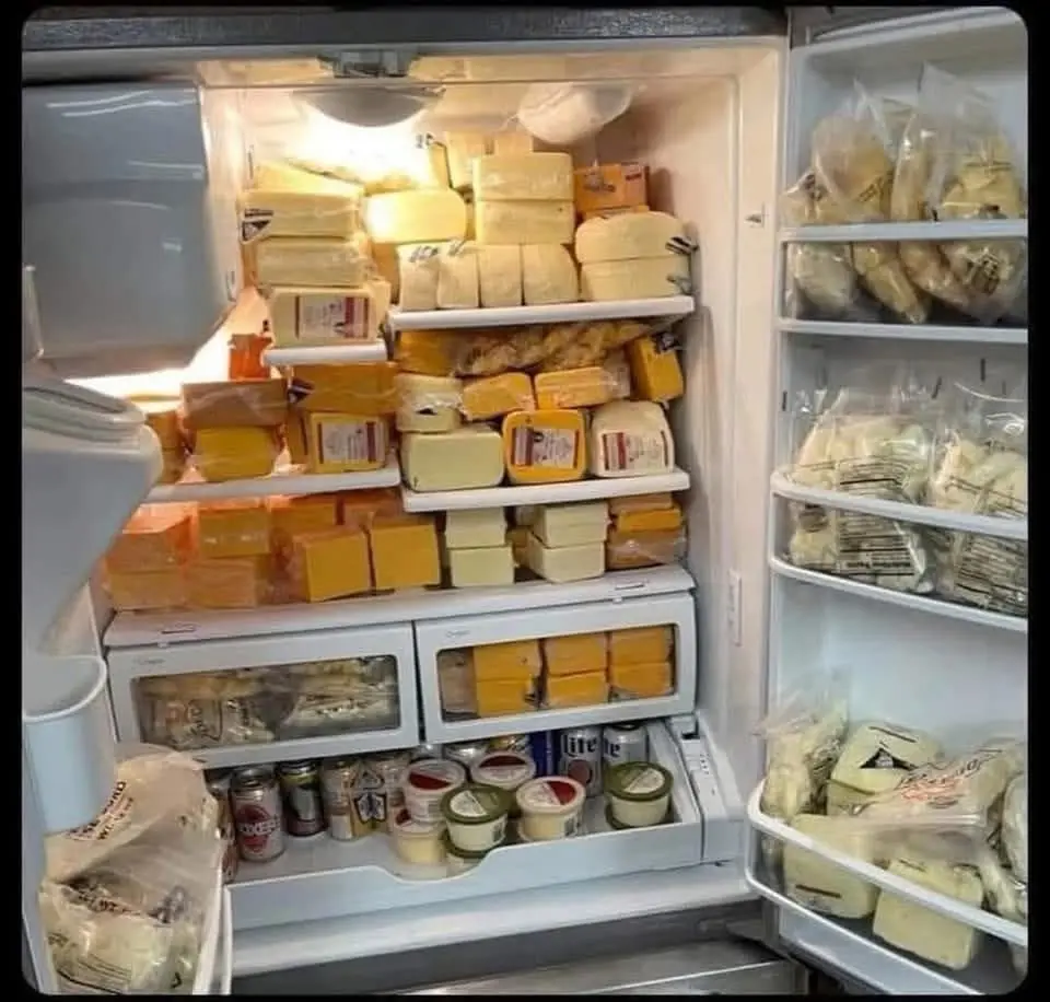 An open refrigerator is completely filled with various types of cheese, stacked on shelves and door compartments, with only…