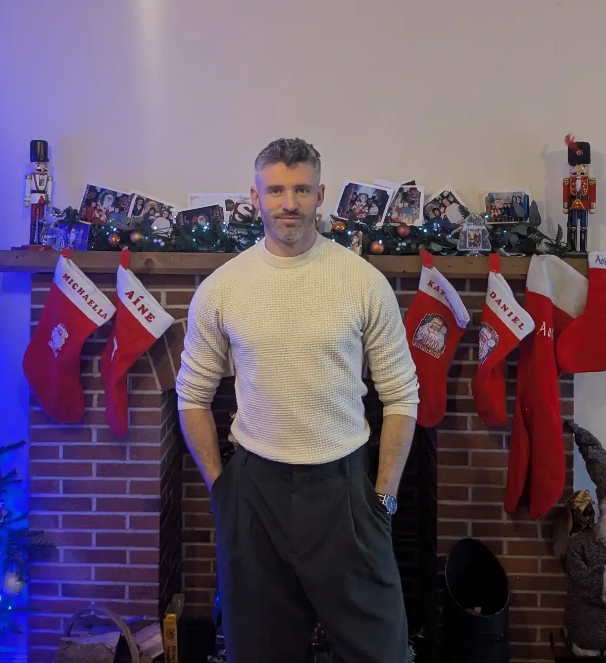 A man stands in front of a fireplace decorated with multiple red Christmas stockings, each labeled with a different name…