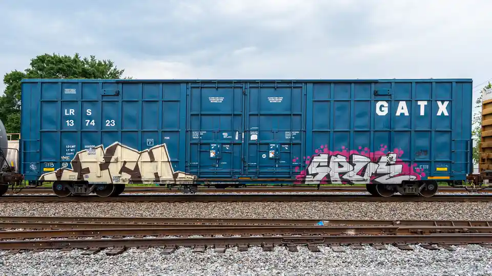 A blue GATX freight train car sits on tracks with graffiti on its side, featuring the text 'gatx' overlaid on the image. The…
