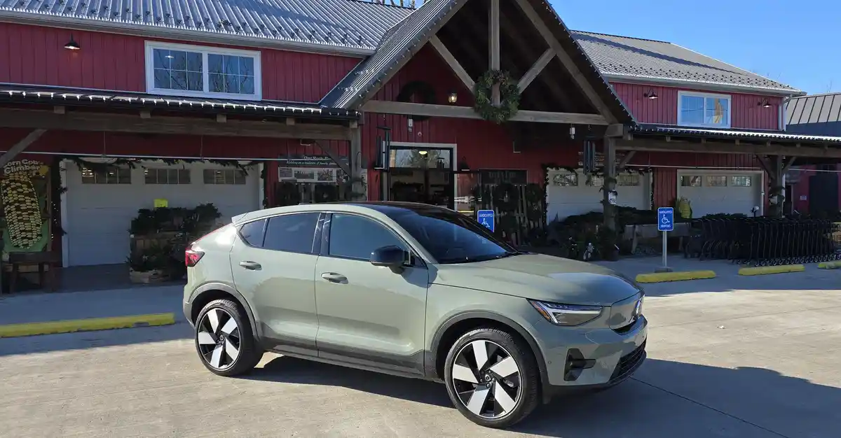 A light green Volvo SUV is parked in front of a red barn-style building decorated with holiday wreaths and garlands. The…