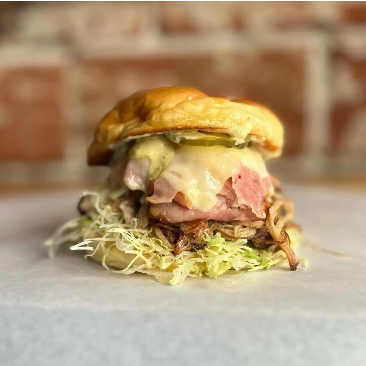 A close-up photo of a gourmet-looking sandwich on a soft bun, layered with melted cheese, slices of pink meat (possibly ham…