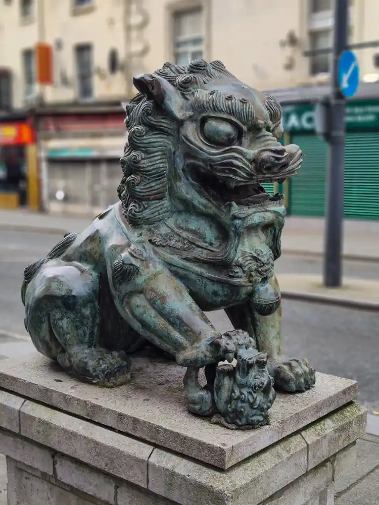 A weathered bronze statue of a mythical lion-like creature, likely a shishi or foo dog, sits crouched on a stone pedestal…