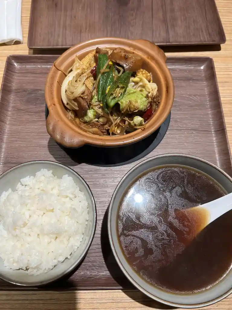 A meal featuring a clay pot of stir-fried vegetables and meat, a bowl of white rice, and a bowl of dark broth with a spoon…
