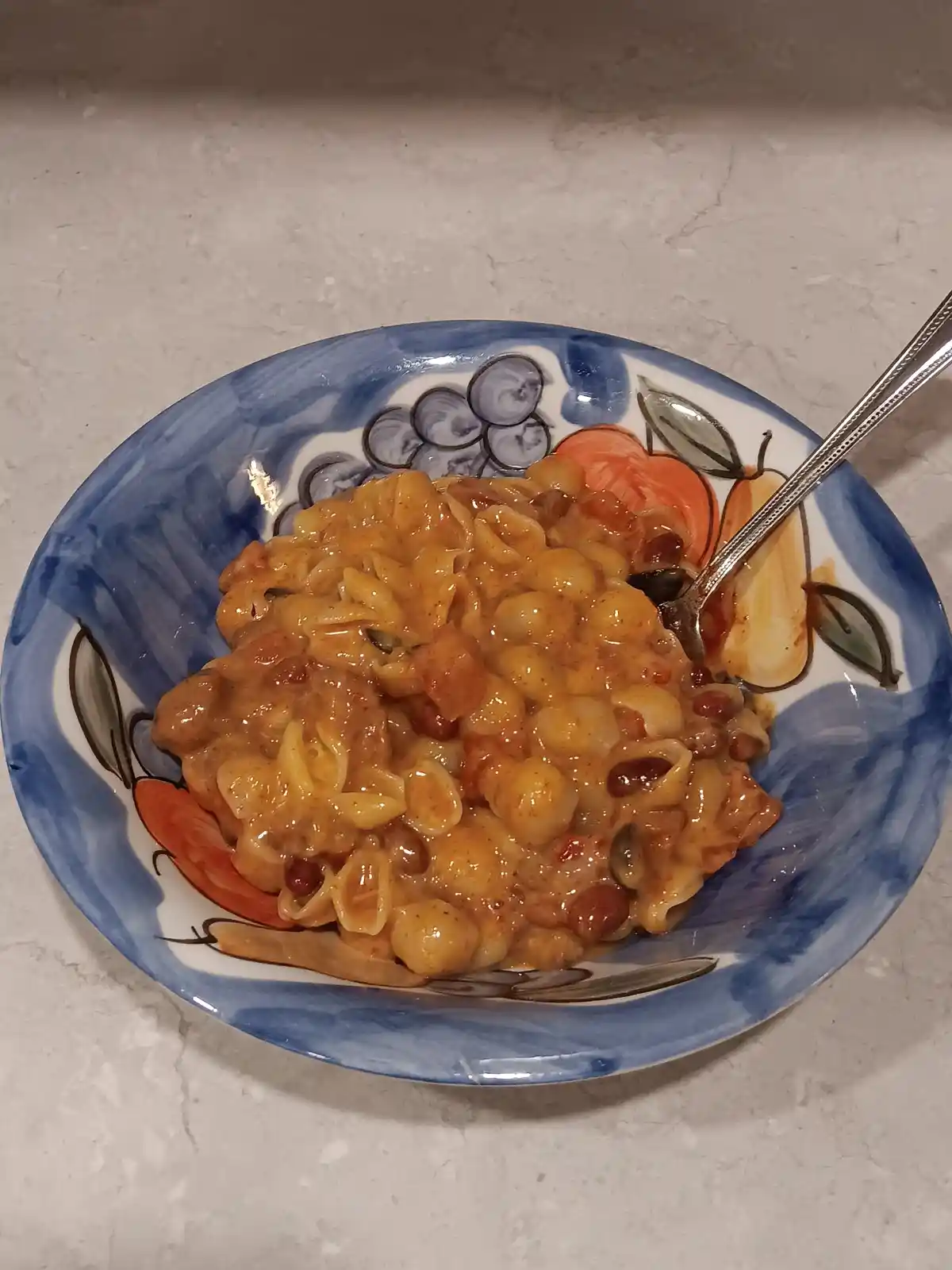 A close-up photo shows a bowl of pasta mixed with beans and a reddish sauce, served in a decorative ceramic bowl with blue…