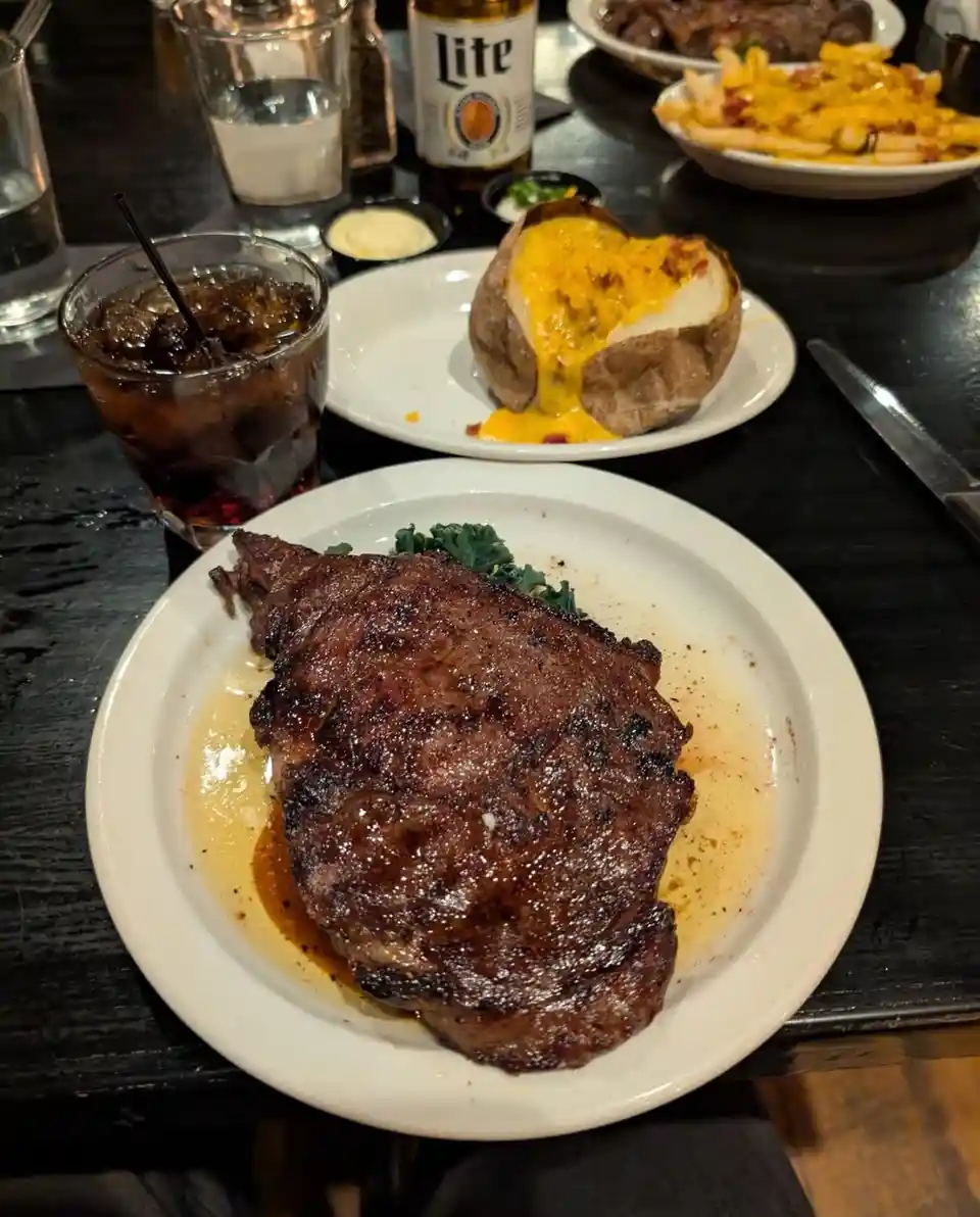 A close-up photo of a large, seared steak on a white plate, accompanied by a side of cheese fries and a baked potato with…
