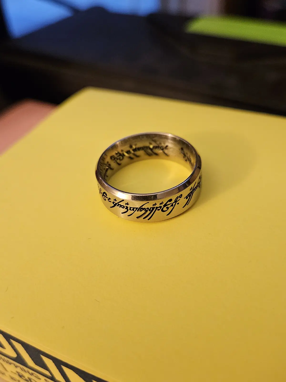 A silver ring engraved with Elvish script from The Lord of the Rings sits on a bright yellow book cover. The overlaid text…