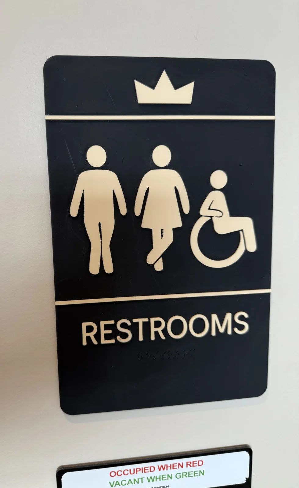 A black restroom sign with gold icons for male, female, and wheelchair-accessible users, topped with a crown, indicating a…