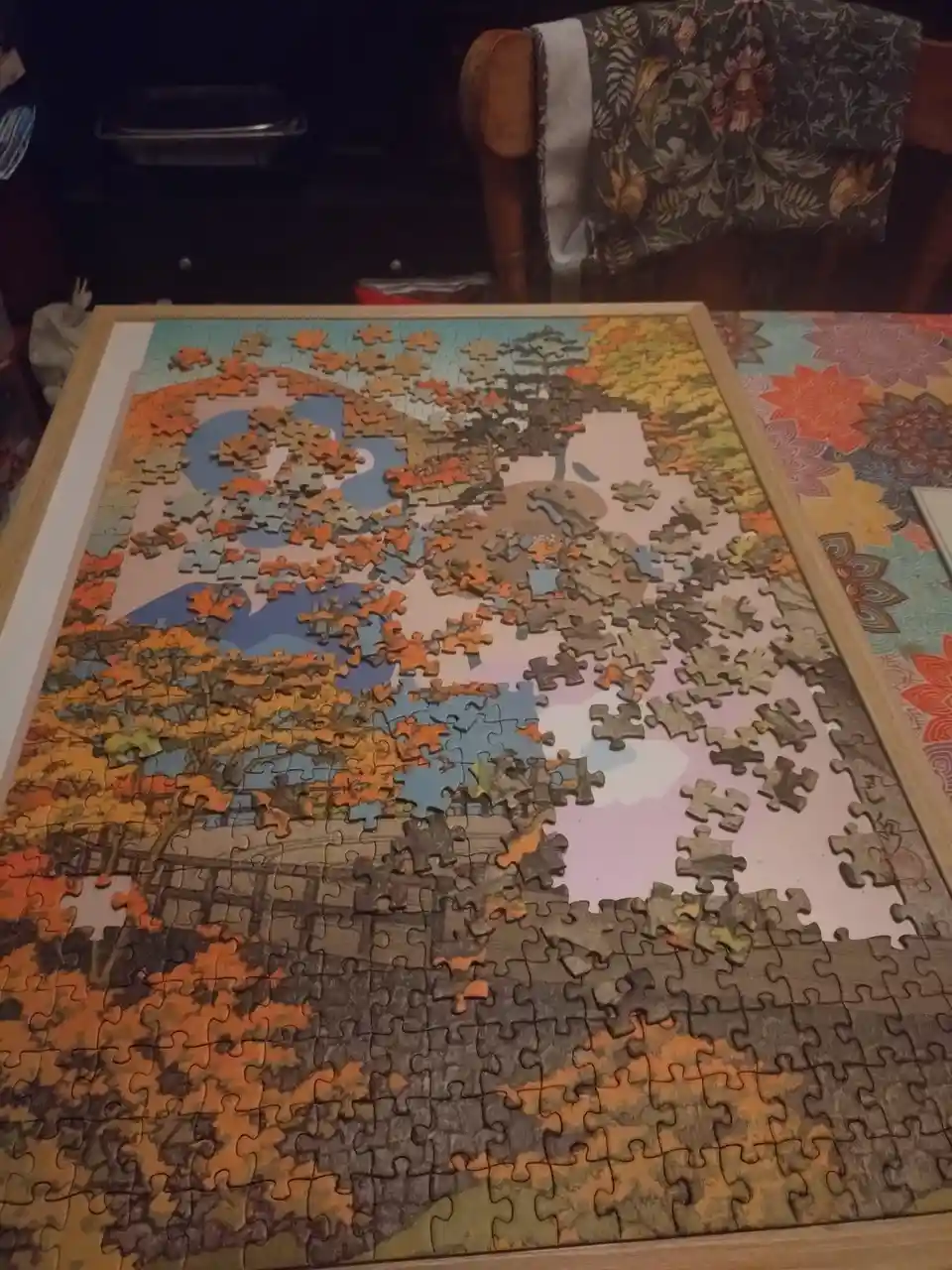 A partially completed jigsaw puzzle depicting autumn trees and a bridge is laid out on a table, with large sections of the…