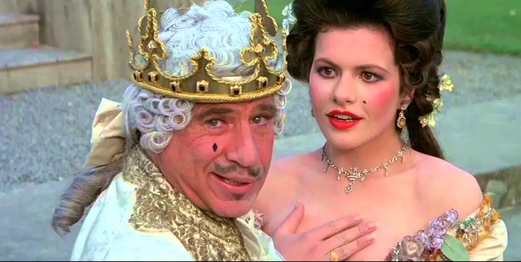 A man with a gold crown and white wig, wearing a mustache and a black dot on his cheek, smiles while a woman in a period…