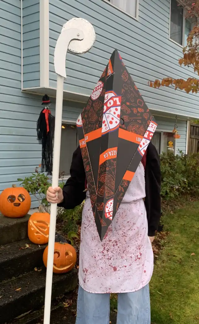 A person dressed in a Halloween costume stands outside a house, wearing a large, pointed hat made from pizza box material, a…