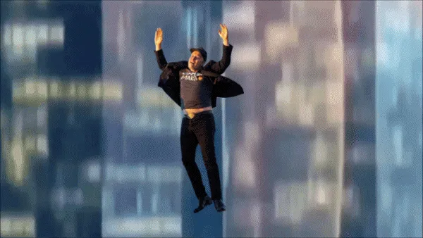 The image shows a man in a dark jacket and pants, arms raised in a joyful or triumphant pose, seemingly floating or falling…