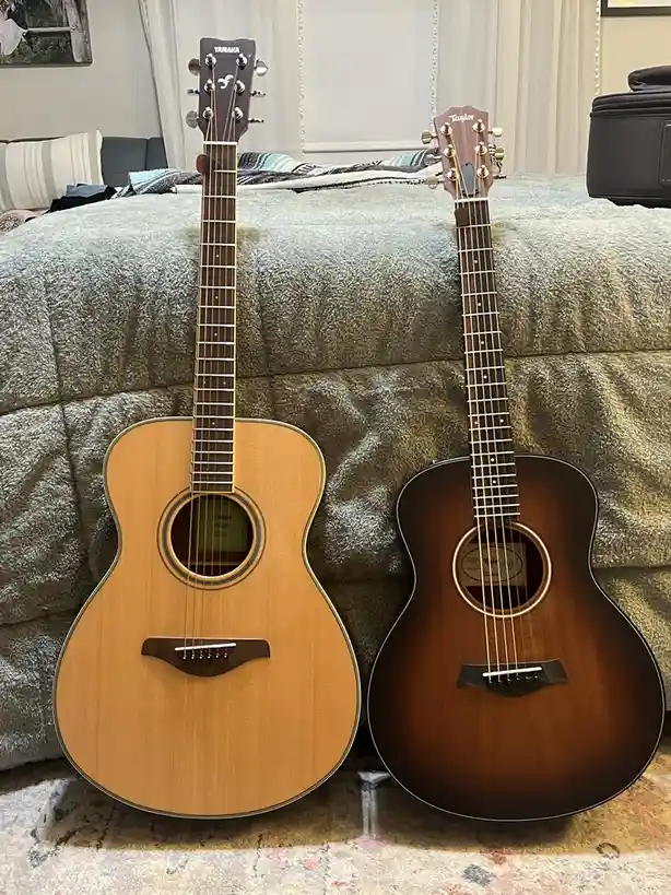 Two acoustic guitars, a light-wood Yamaha and a dark-wood Taylor, stand side-by-side on a textured gray couch in a living…