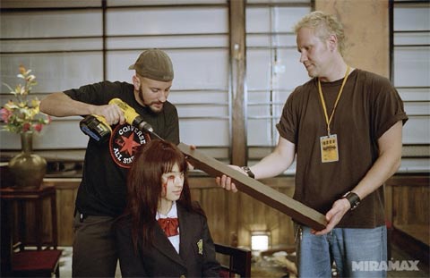 A man in a black t-shirt with a 'Don't Cut the Hair' logo uses a power drill to cut the hair of a seated woman in a school…