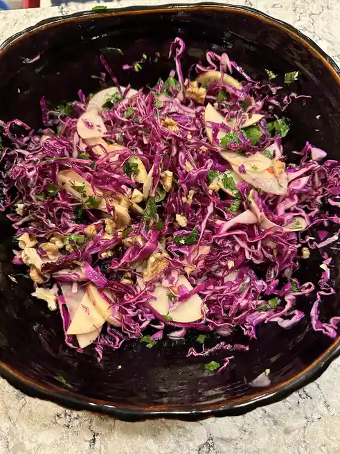 A close-up photo shows a dark bowl filled with a colorful salad made of shredded purple cabbage, sliced apples, chopped…
