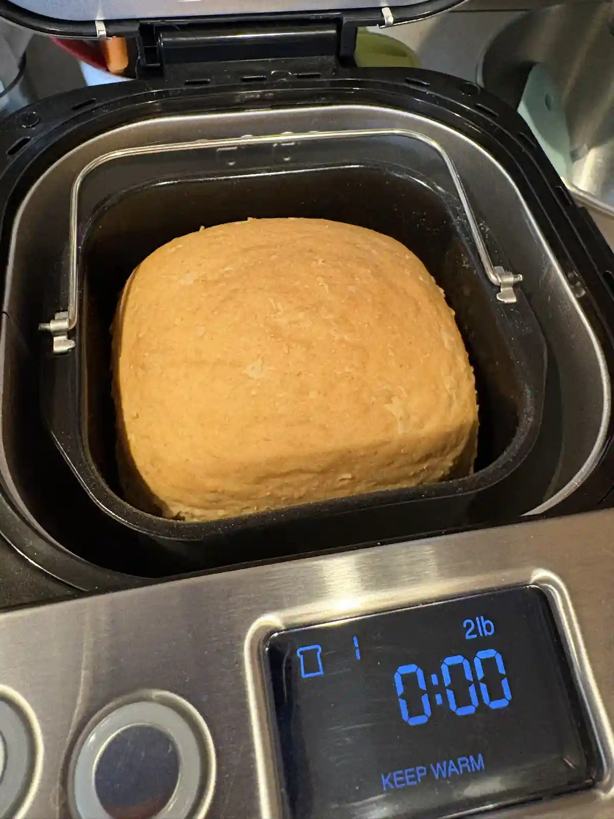 A freshly baked loaf of bread sits inside a bread machine, which displays '0:00' and '2lb' on its digital screen with 'KEEP…