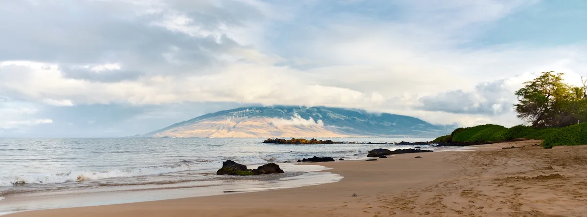 A serene beach panorama with gentle waves, sandy shore, and a distant mountain range partially obscured by clouds. The…