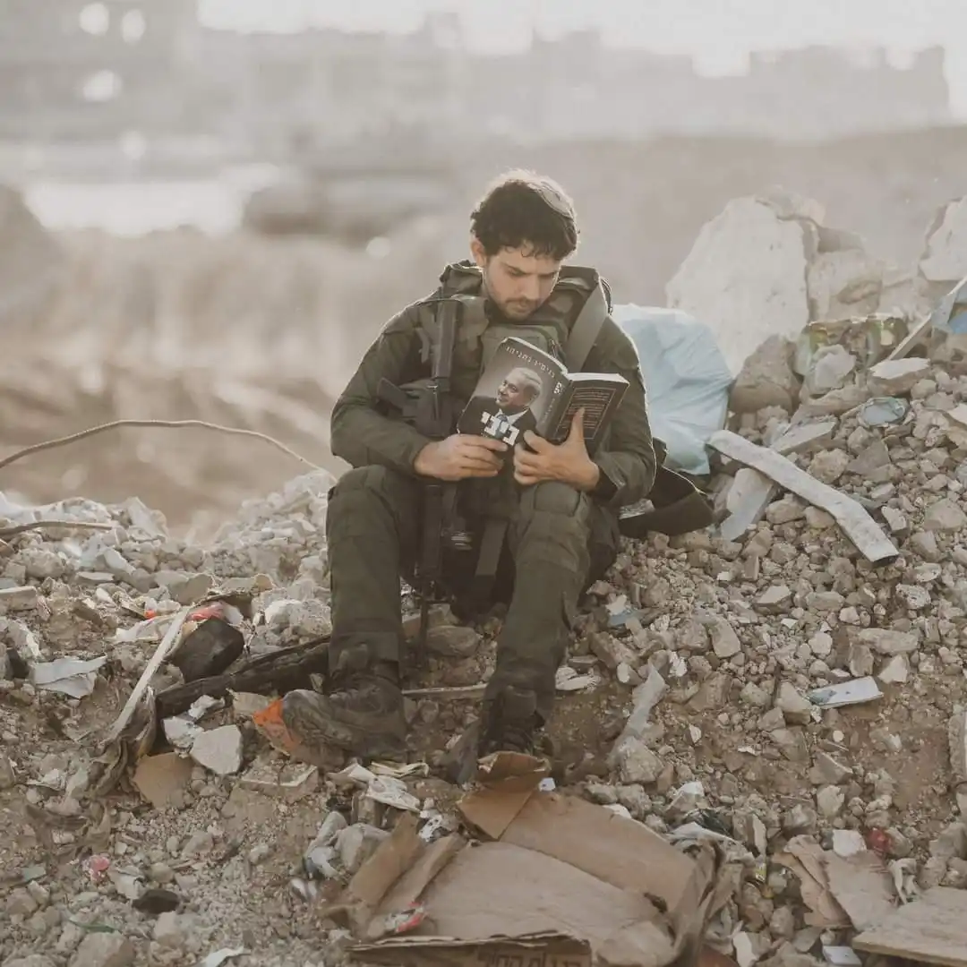 A soldier in military gear sits amidst rubble and debris, holding an open book with a photo of Israeli president Isaac…