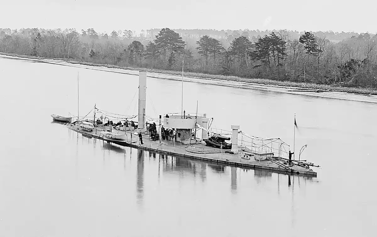 A black-and-white historical photograph shows a flat, open-decked steamboat on a calm river, with trees lining the distant…