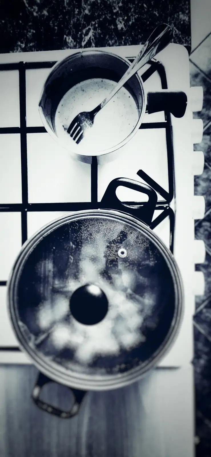 A black and white overhead shot of a kitchen stove with two pots. The top pot contains a white liquid with a fork resting…