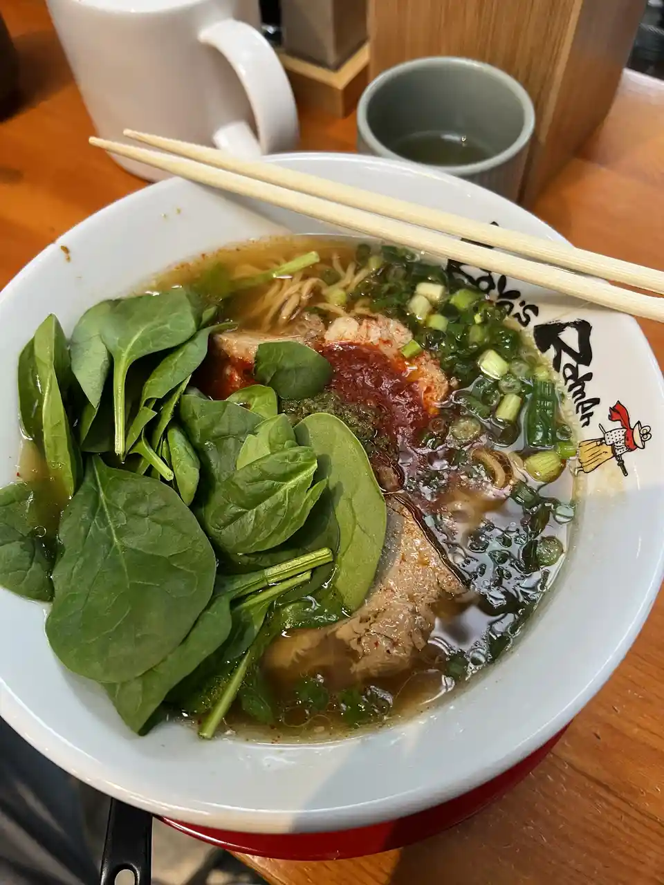 A close-up photo shows a white ramen bowl filled with broth, noodles, slices of meat, a large pile of fresh spinach, chopped…