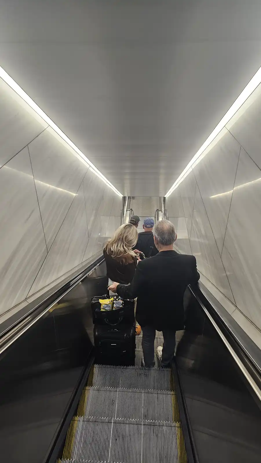 A person with gray hair and a black jacket is riding an escalator upward, pulling two black suitcases. The top suitcase has…