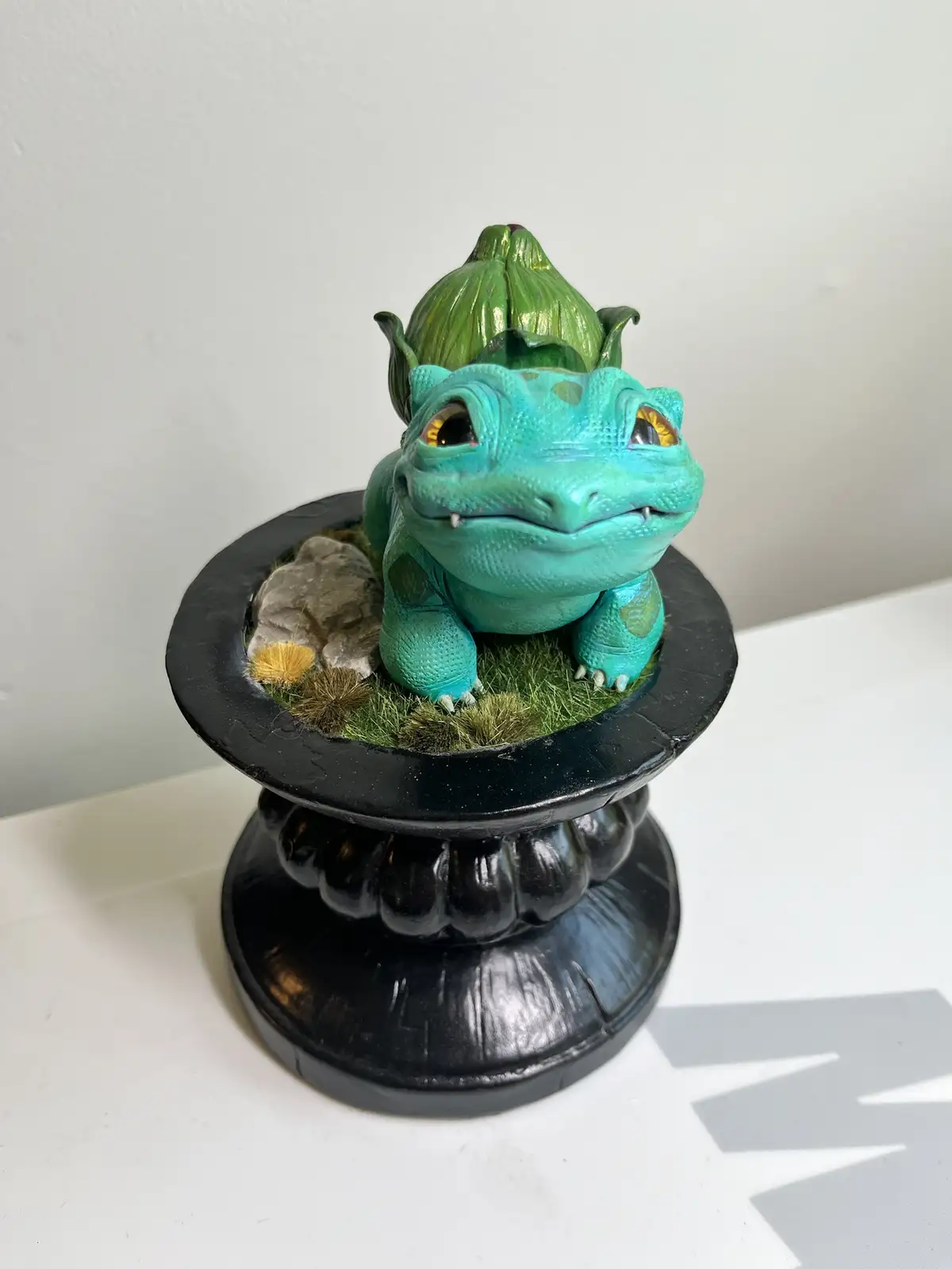 A turquoise, cartoonish dragon figurine with a green artichoke-like crown sits in a black decorative planter filled with…