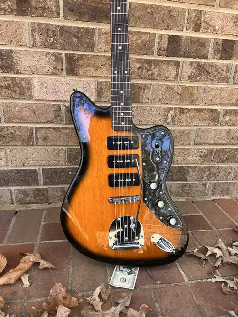 A sunburst-finish electric guitar with a black pickguard and chrome hardware leans against a brick wall on a brick patio…
